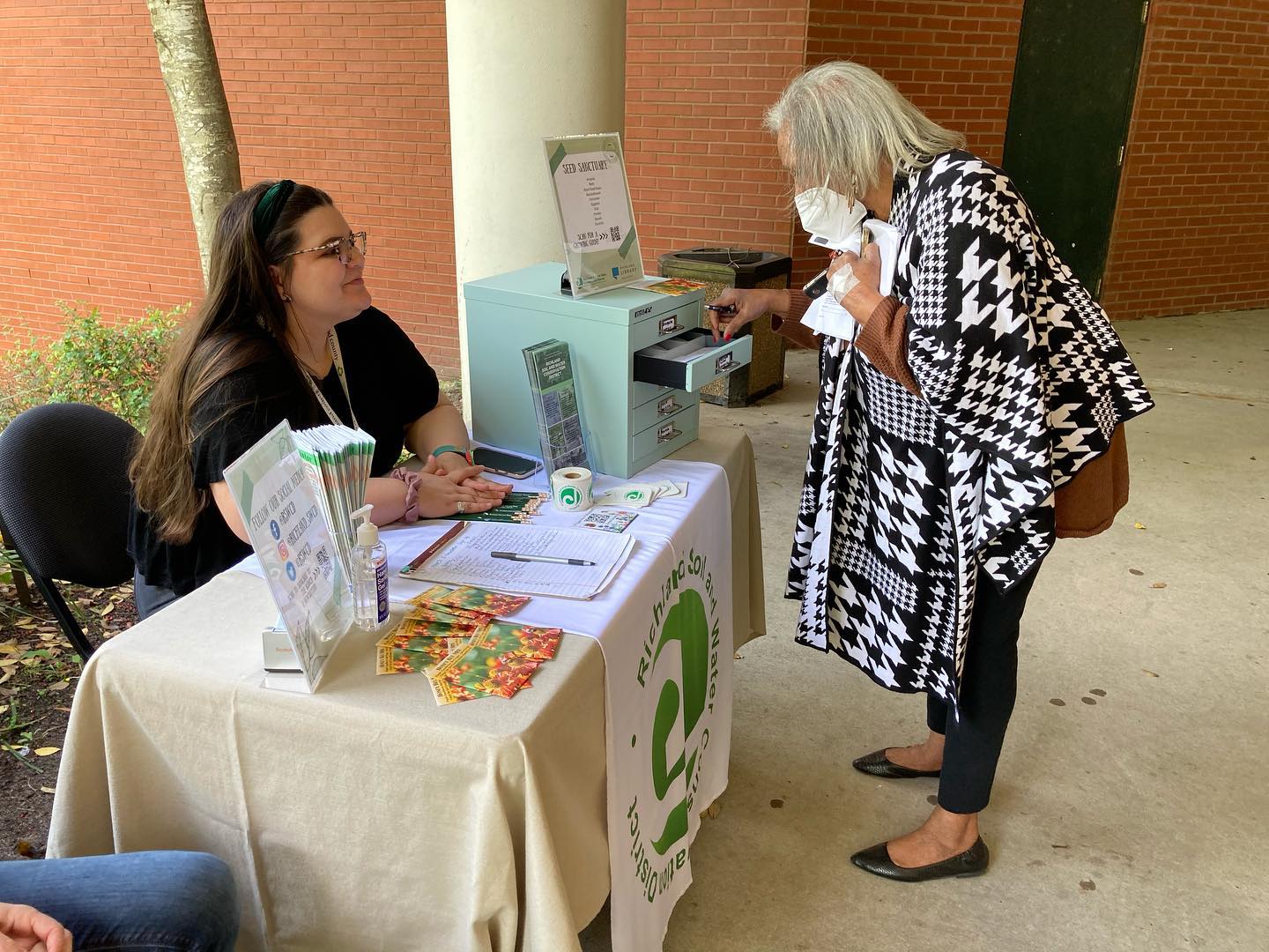 A Richland Soil and Water Conservation District employee sits behind a table at a community event while a member of the public takes seeds from the Seed Sanctuary.